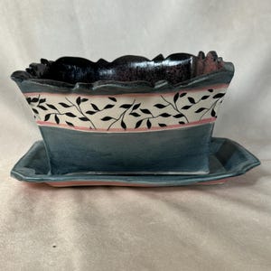 Puede incluir: Una maceta de cerámica azul y rosa con un diseño de hojas blancas y negras. La maceta tiene un platillo a juego.