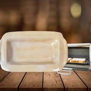 以下が含まれることがあります： わずかに盛り上がった縁のある白いセラミック製の長方形のベーキングディッシュ。ディッシュは、トースターオーブンと2枚のパンが入った木の表面の上に置かれています。
