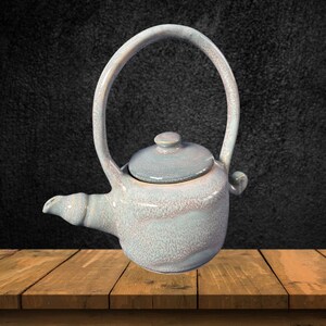 May include: A light blue ceramic teapot with a textured finish and a handle that forms a loop. The teapot has a lid and a spout.