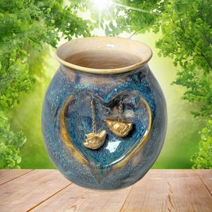 Op de afbeelding: Een blauwe keramische vaas met een hartvormig ontwerp en gouden accenten. Twee goudkleurige decoratieve elementen hangen aan het hart. De vaas heeft een lichtbruine rand en staat op een houten oppervlak.