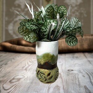 May include: A ceramic vase with a white top, a green and brown middle section, and a light green base featuring intricate cut-out floral patterns. The vase is filled with a lush, leafy green and white plant with patterned foliage.