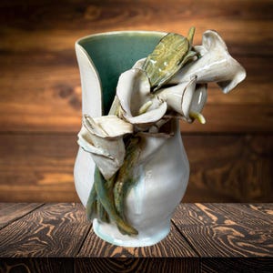 Peut inclure: Un vase en céramique blanc avec un intérieur turquoise. Le vase est orné de callas blancs sculptés et de feuilles vertes. Le vase est présenté sur une surface en bois.