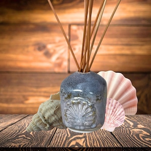 May include: A ceramic reed diffuser bottle with a mottled blue and grey glaze, featuring a raised shell relief on the front. Several thin wooden reeds extend from the top. The bottle is flanked by a textured beige conch shell and a pink fan shell.