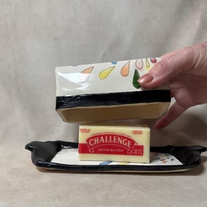 Puede incluir: Un mantequillero de cerámica blanca con un borde negro y un diseño de gotas de lluvia de colores. El mantequillero está abierto y contiene una barra de mantequilla salada Challenge. El mantequillero está sobre una bandeja de cerámica negra y blanca con un diseño similar de gotas de lluvia.
