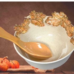 May include: A ceramic serving bowl with a cream interior and white exterior. The rim is decorated with gold oak leaves. A matching spoon rests inside the bowl. Pumpkins are in the foreground, suggesting an autumnal theme.