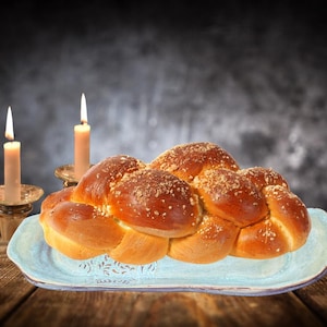 Puede incluir: Un pan challah trenzado con una ligera capa de azúcar en la parte superior, descansando sobre una fuente de cerámica azul y blanca. Dos velas encendidas están en el fondo.