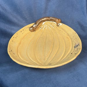 Könnte beinhalten: Eine gelbe Keramik-Servierplatte in Form eines Kürbisses mit einem braunen Griff. Die Platte hat eine strukturierte Oberfläche und schwarze Flecken.