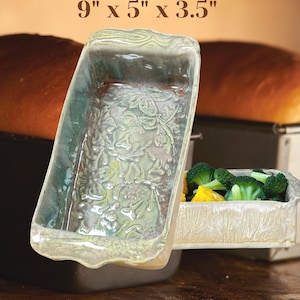 May include: A rectangular ceramic loaf pan with a sage green glaze and a raised floral relief pattern on the interior base. The exterior features a wood grain texture. The dimensions 9" x 5" x 3.5" are printed in brown text above the object.