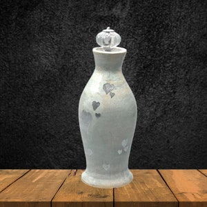 Puede incluir: Un jarrón de cerámica azul claro con un tapón de vidrio transparente y un diseño de corazones plateados. El jarrón está sobre una superficie de madera.