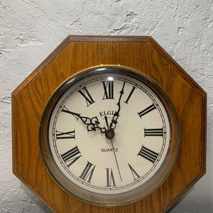 May include: Octagonal wooden wall clock with a cream-colored face and black Roman numerals. The clock face has the brand name "ELGIN" and the word "QUARTZ" printed on it. The clock hands are black.