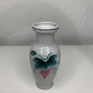 May include: A small white ceramic vase with a gold rim and a painted design of pink grapes and green leaves.