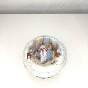 May include: A white porcelain trinket box with a round lid. The lid features a colorful painted scene of people dancing in a room. The box is decorated with a white and pink floral design around the edge.
