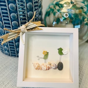 Puede incluir: Una imagen enmarcada en blanco con una pieza de arte de pájaros de conchas marinas y vidrio de mar. Los pájaros son verdes y amarillos y están posados ​​en una roca y conchas marinas.