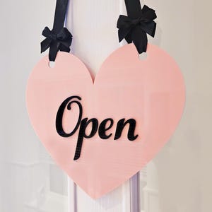 May include: A pink heart-shaped sign with black ribbon bows and the word "Open" in black cursive lettering.