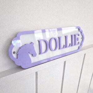 May include: A rectangular wall sign with rounded, decorative edges featuring a light purple border and a white centre. The sign displays the name DOLLIE in raised light purple capital letters next to a silhouette of a horse head in light purple.
