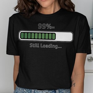 May include: Black t-shirt featuring a white graphic of a loading bar at 99% and the words "Still Loading...". The shirt has a crew neck and is made of a soft material.