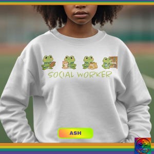 Puede incluir: Una sudadera blanca con un gráfico verde de cuatro ranas de dibujos animados, cada una realizando diferentes actividades. El texto "SOCIAL WORKER" está impreso en verde debajo. La palabra "ASH" está en un rectángulo de color en la parte inferior.