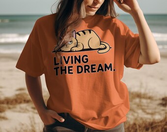 May include: Orange t-shirt featuring a cartoon cat illustration with the text "LIVING THE DREAM." The cat is light brown with dark brown stripes. The text is in a bold, black font. The shirt is a solid orange color.