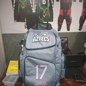 May include: A grey rolling backpack with the word "AZTECS" in teal and red. The number "17" in teal and the name "GARZA" in pink are on the front. Jerseys with deer graphics and the name "AMAZO" are on the wall behind the backpack.