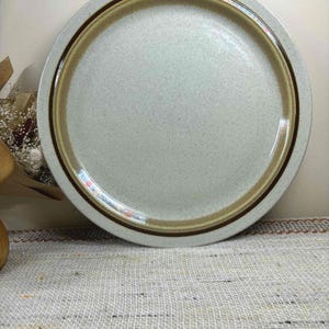May include: A round, speckled, light grey ceramic plate with a brown and tan rim. The plate has a simple, minimalist design, suitable for serving food. The plate is on a textured, light-coloured surface.