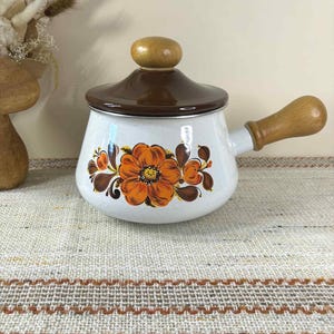 Puede incluir: Cacerola de esmalte blanco con tapa marrón y asa de madera. La olla presenta un gran diseño floral naranja con hojas marrones. La tapa tiene un pomo de madera. La cacerola está sobre una superficie texturizada de color neutro.