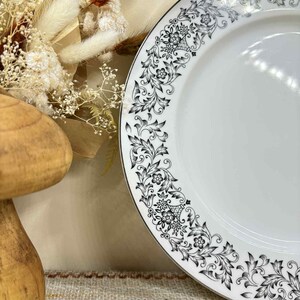 Puede incluir: Plato de cerámica blanco con un patrón floral negro alrededor del borde. El plato tiene un borde plateado y un diseño simétrico detallado. El plato es parte de un juego de vajilla, perfecto para servir comidas.