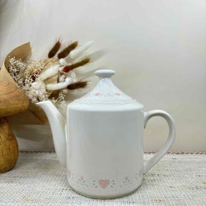 May include: White ceramic teapot with a lid, decorated with a pink heart and floral pattern around the base and lid. The teapot has a curved spout and a handle. A bouquet of dried flowers is in the background.
