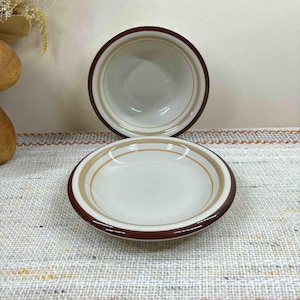 Puede incluir: Dos cuencos de cerámica blanquecina con borde marrón y diseño de rayas beige y marrón. Los cuencos están vacíos y se encuentran sobre una superficie estampada. Los cuencos son probablemente para servir alimentos.
