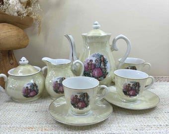 May include: A vintage tea set with a pale yellow finish. The set includes a teapot, sugar bowl, creamer, and cups with saucers. Each piece features a decorative image of a couple in period clothing. The set is arranged on a textured surface.