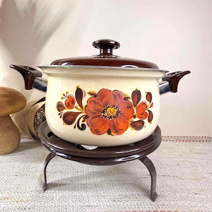 May include: Cream enamel pot with a brown lid and handles, decorated with a large orange floral design. The pot is on a dark brown metal trivet. A vintage kitchenware item.