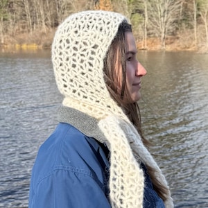 May include: A cream-colored, open-weave hood and scarf set. The hood is worn over a blue jacket, with the scarf draped over the shoulders. The background features a lake and trees, suggesting outdoor wear.