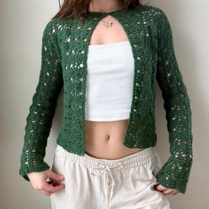 以下が含まれることがあります： ダークグリーンのオープンフロントのかぎ針編みカーディガン、長袖。カーディガンは繊細な透かし模様のデザインです。白いチューブトップとベージュのドローストリングパンツを着用しています。金のネックレスが見えます。