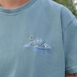 Puede incluir: Una camiseta de color verde azulado con un bordado de montaña y sol. El diseño presenta una cordillera blanca, un sol amarillo y un río azul. La camiseta está hecha de material suave.