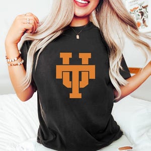 May include: Black t-shirt with a burnt orange graphic of the letters "TT" stacked on top of each other. The shirt is a crew neck style and appears to be made of a soft material. The graphic is centered on the front of the shirt.