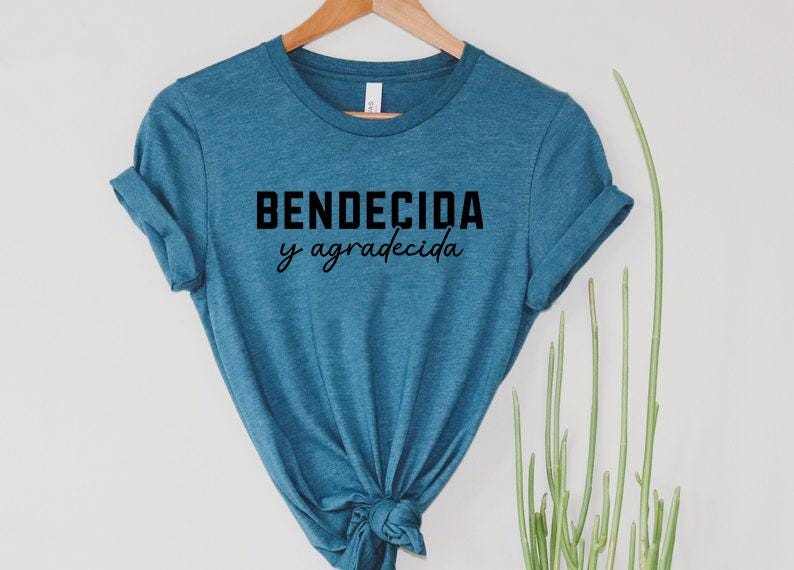 May include: A teal t-shirt with the words "BENDECIDA y agradecida" in black block and script font. The shirt is hanging on a wooden hanger against a white background, with green plants to the side.