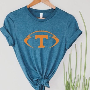 May include: A teal-colored t-shirt with a large orange football design featuring the letter "T" in the center. The shirt is hanging on a wooden hanger against a white background. The shirt is knotted at the bottom.