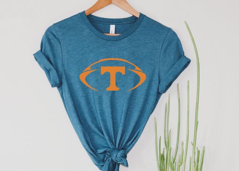 May include: A teal-colored t-shirt with a large orange football design featuring the letter "T" in the center. The shirt is hanging on a wooden hanger against a white background. The shirt is knotted at the bottom.