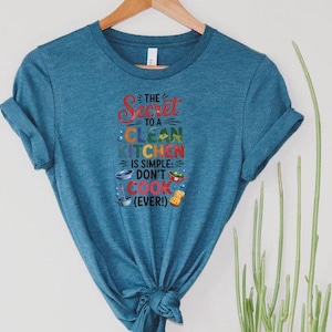 May include: A teal t-shirt with the text "The Secret to a Clean Kitchen is Simple: Don't Cook (Ever!)" in colorful lettering. The shirt features a crew neck and rolled-up sleeves. The shirt is hanging on a wooden hanger.