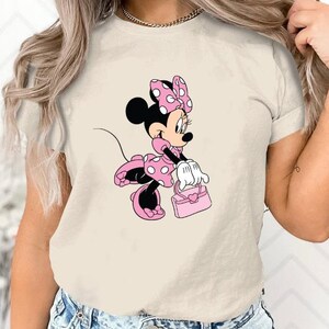 May include: A beige t-shirt with a cartoon image of Minnie Mouse wearing a pink dress with white polka dots and a pink bow. She is holding a pink purse.