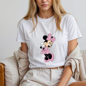 May include: White t-shirt with a pink and black Minnie Mouse graphic. Minnie Mouse is wearing a pink bow and a pink polka dot dress.