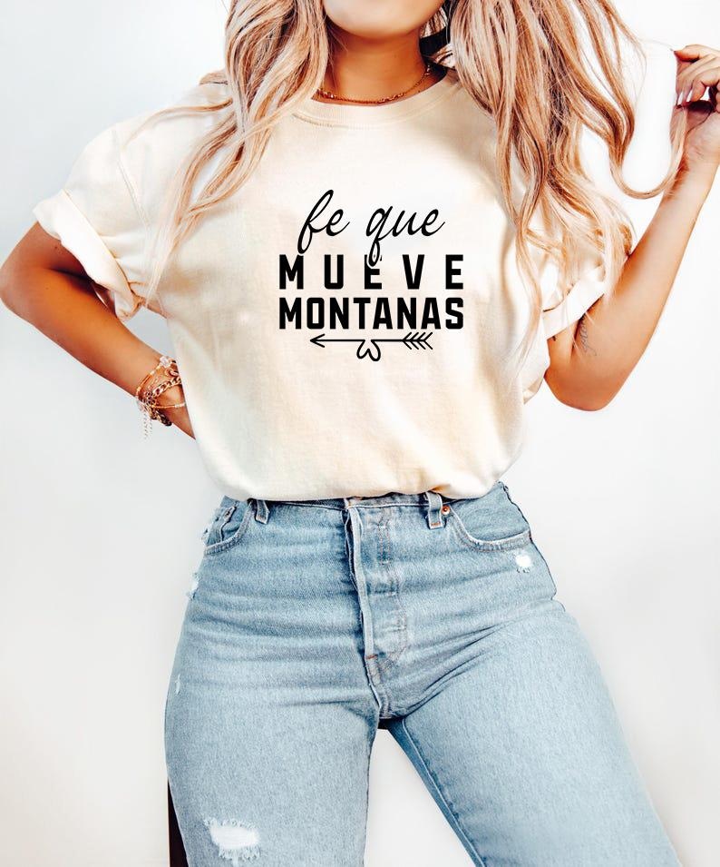 May include: Cream-colored t-shirt with the Spanish text "fe que mueve monta&ntilde;as" in black, meaning "faith that moves mountains." The shirt is paired with light blue denim jeans. The text is centered and includes an arrow design.