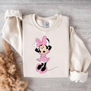 May include: A cream-colored sweatshirt with a graphic of Minnie Mouse wearing a pink dress with white polka dots and a pink bow.