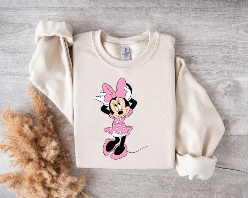 May include: A cream-colored sweatshirt with a graphic of Minnie Mouse wearing a pink dress with white polka dots and a pink bow.