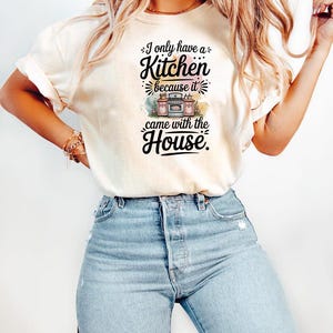 May include: Cream-colored t-shirt with the text "I only have a Kitchen because it came with the House." The text is in black, with a colorful illustration of a kitchen oven and cabinets. The shirt is paired with light blue jeans.