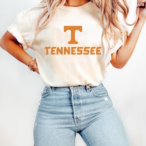 May include: Cream-colored t-shirt with the word "TENNESSEE" in orange block letters. The letter "T" is above the word "TENNESSEE". The shirt is paired with light blue denim jeans.