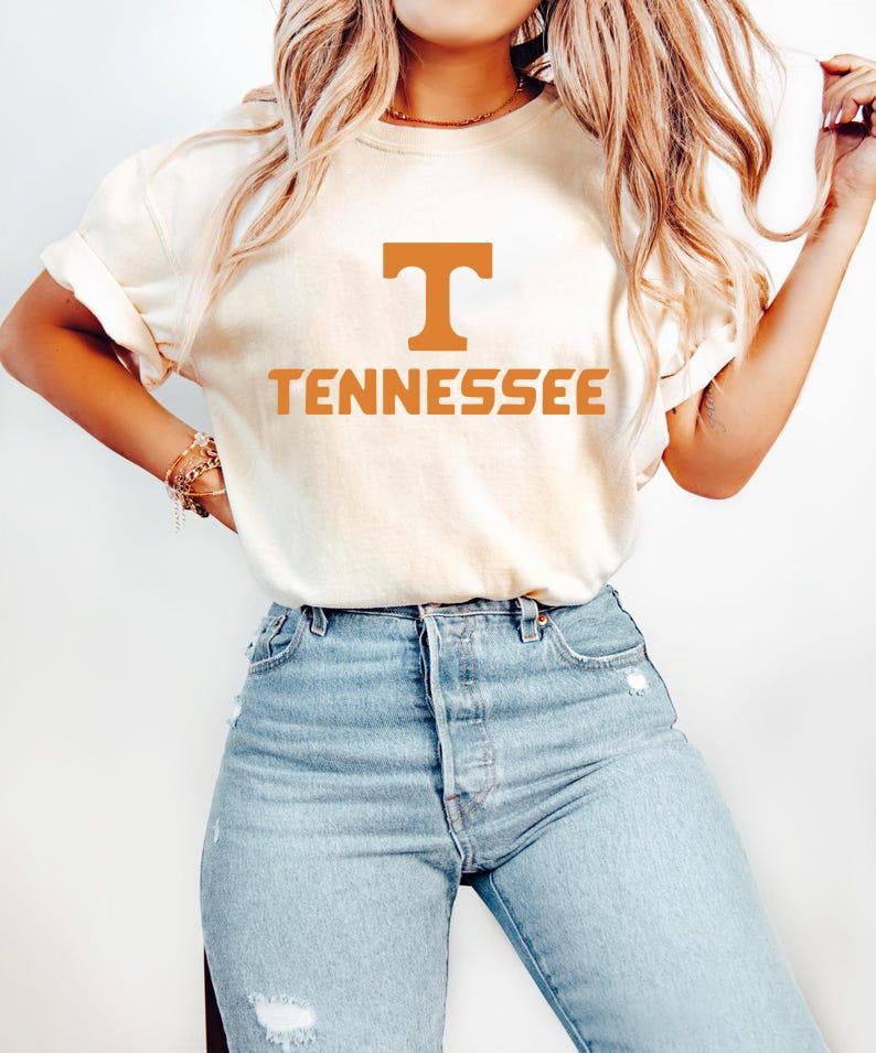 May include: Cream-colored t-shirt with the word "TENNESSEE" in orange block letters. The letter "T" is above the word "TENNESSEE". The shirt is paired with light blue denim jeans.
