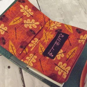 May include: A small green and teal fabric journal with a zipper closure and a red strap. The cover is decorated with a colorful autumn leaf pattern and the text "Little Bits" in pink.