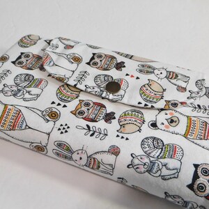May include: A white fabric wallet with a flap closure and a snap button. The wallet features a repeating pattern of owls, bears, rabbits, and birds with geometric designs in shades of orange, yellow, green, and red.