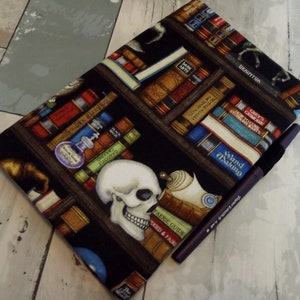 May include: A black fabric book cover featuring a bookshelf design with books, potions, and a skull. The cover has a pen resting on it. The books have titles like "Wand Making" and "Faerie Guide".