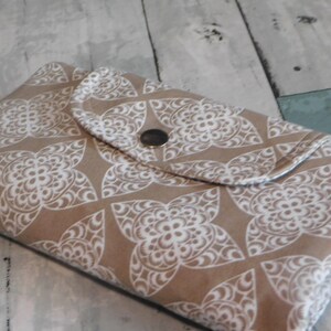 May include: A rectangular fabric pouch featuring a tan background with a repeating white geometric floral pattern. The pouch has a fold-over flap secured by a single centered silver-toned metal snap button closure, creating a flat, envelope-style design.